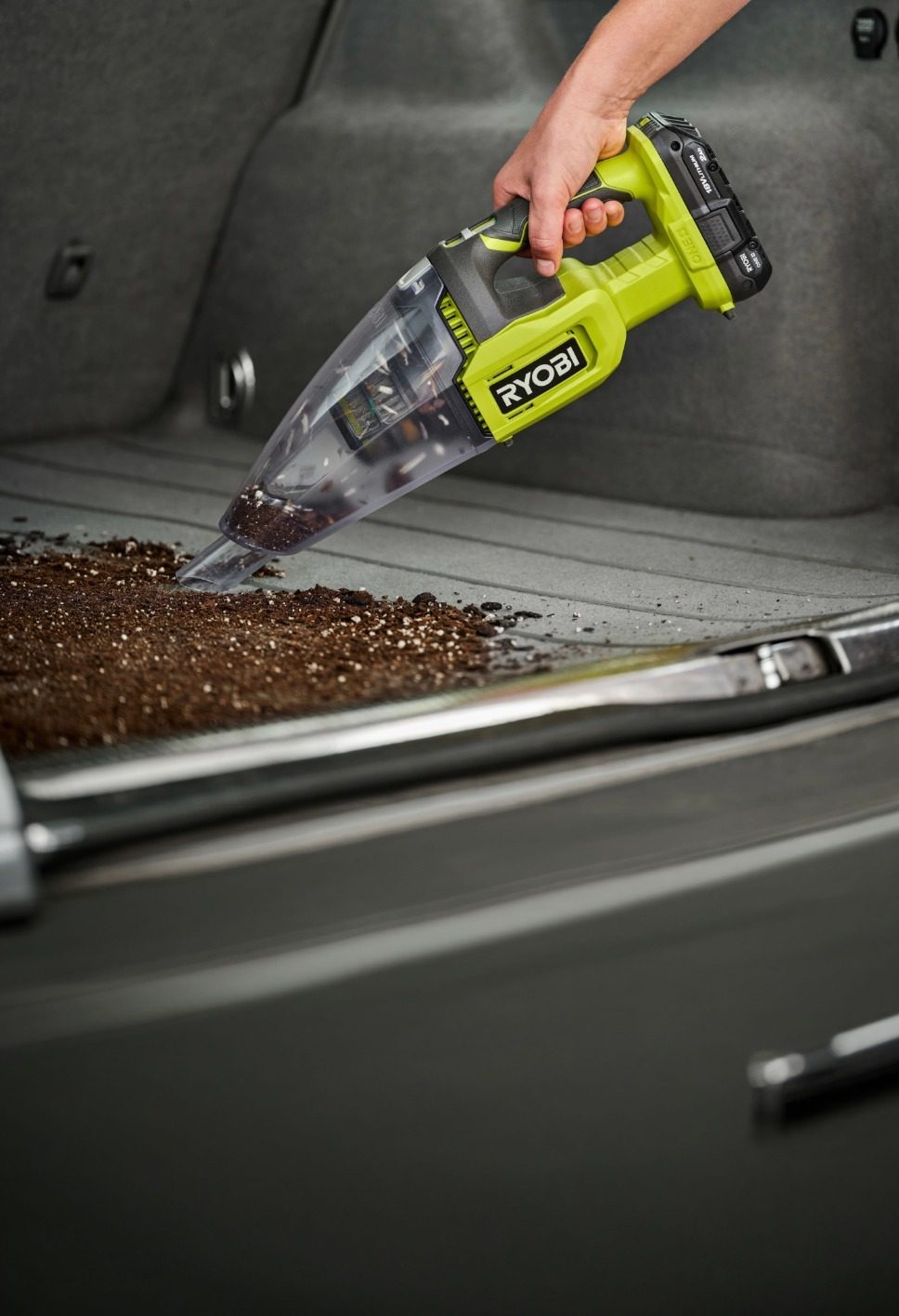 A hand holding a bright green Ryobi cordless vacuum cleaner removes dirt from a car's trunk.