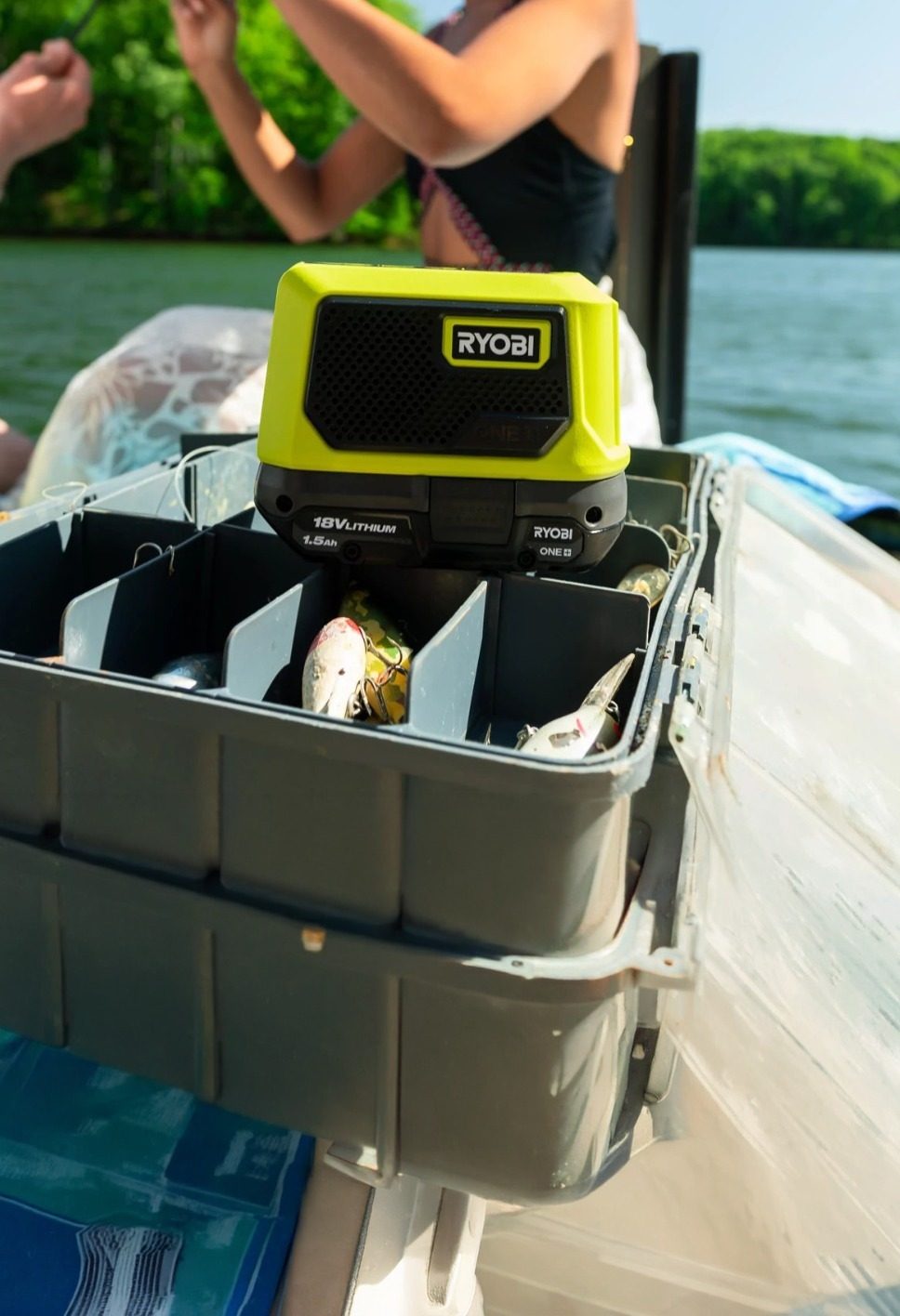 A lime green Ryobi radio sits on a tackle box filled with fishing lures on a boat by a lake.