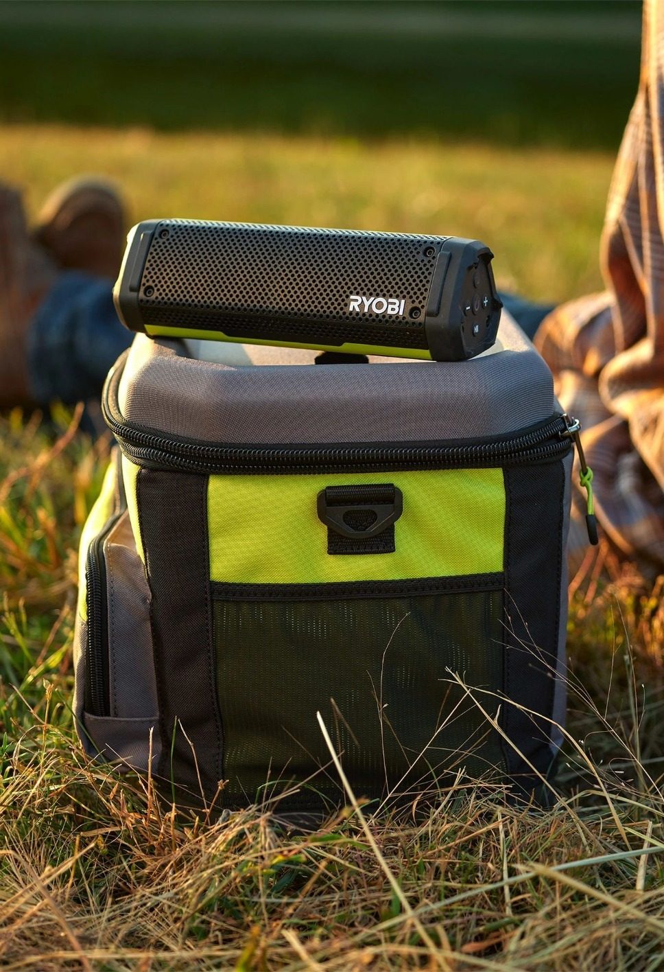Ryobi portable speaker resting on a matching lime green and gray cooler bag in a grassy outdoor setting.