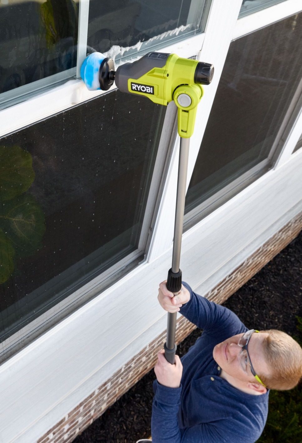 A person uses a long-reach RYOBI power scrubber to clean the exterior of a window.
