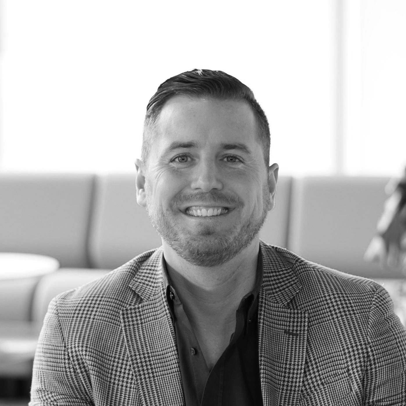 Black and white headshot of a smiling man with a beard, wearing a plaid blazer, looking at the camera.