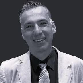 Smiling man in a light suit jacket, dark shirt, and striped tie against a dark background.