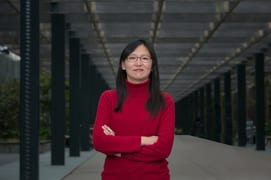 A woman in a red turtleneck and glasses stands with arms crossed in a modern outdoor corridor.