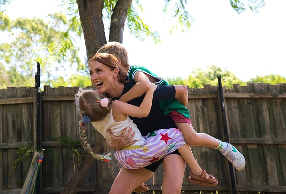 Smiling woman piggybacking two children, a girl and a boy, in a sunny backyard.
