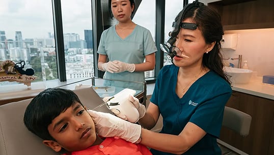 A medical professional examining a child's ear with specialized equipment, assisted by another staff member.
