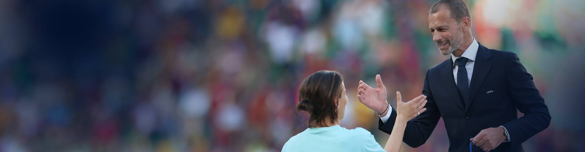 Smiling man in UEFA suit and woman extending hands at stadium.