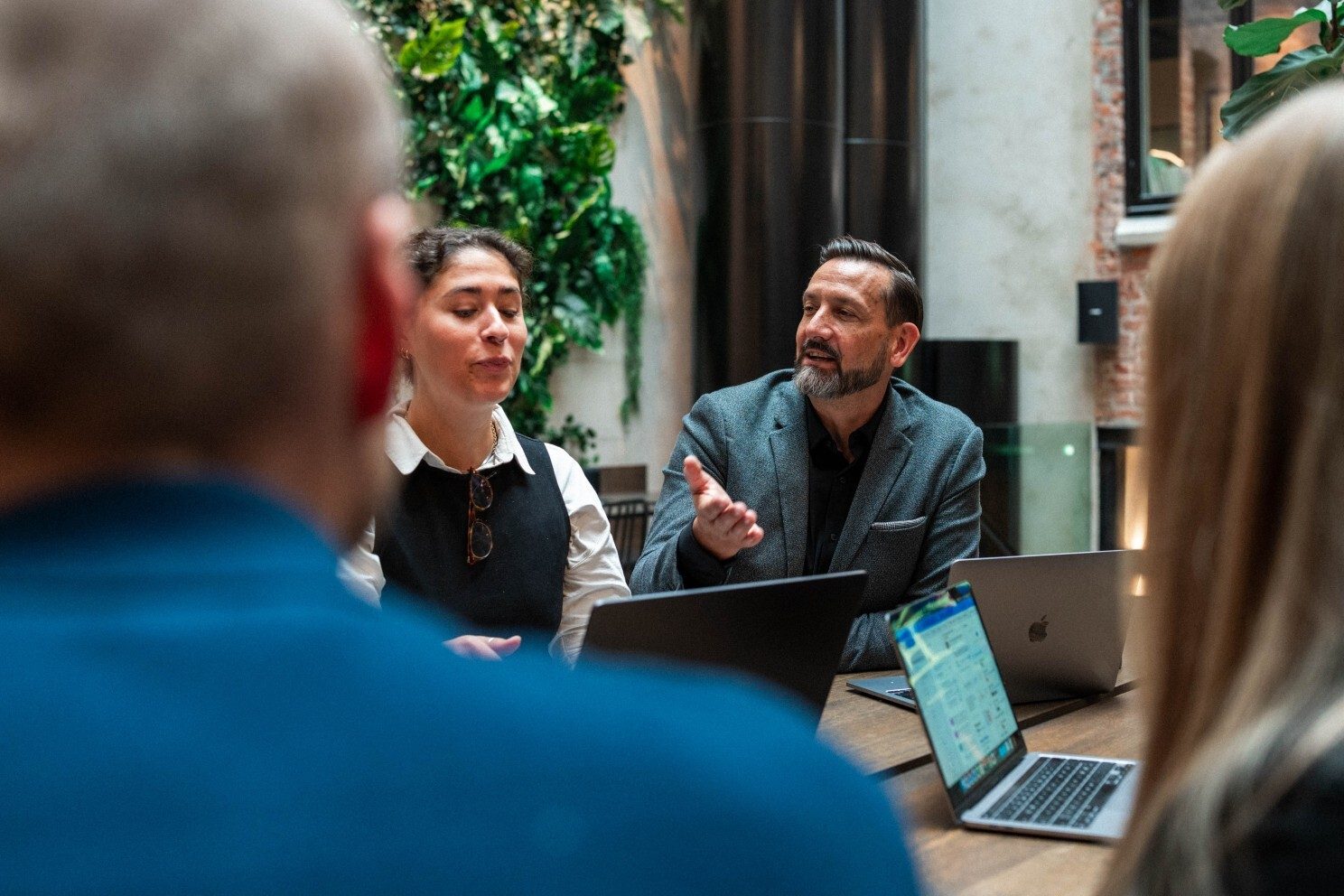 A man speaks, gesturing, at a meeting with a woman, other attendees, and laptops.
