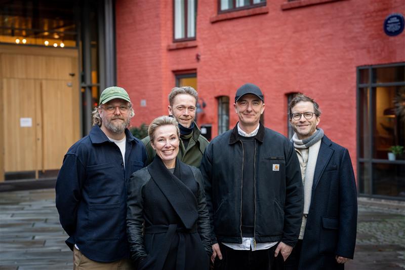 Four men and one woman smiling, standing outdoors in front of a red building.