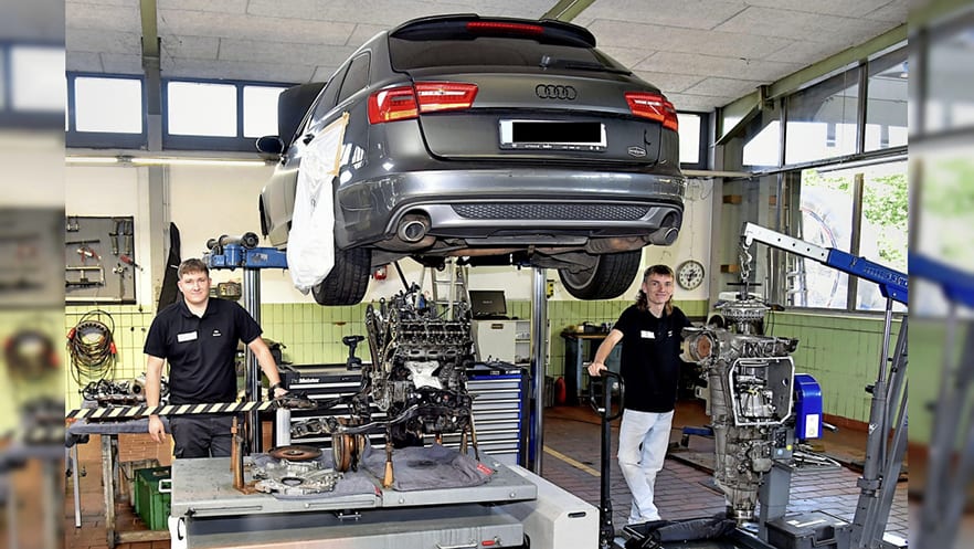 Two mechanics in a garage, one standing by a lifted Audi wagon, the other near disassembled engines.
