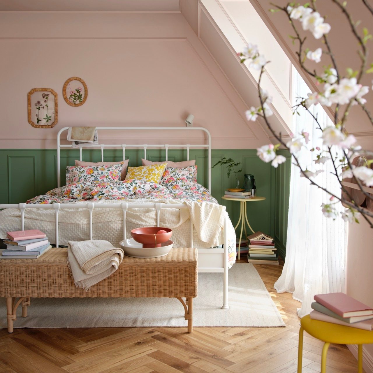 A cozy bedroom with pink & green walls, white bed, floral bedding, and a wicker bench.