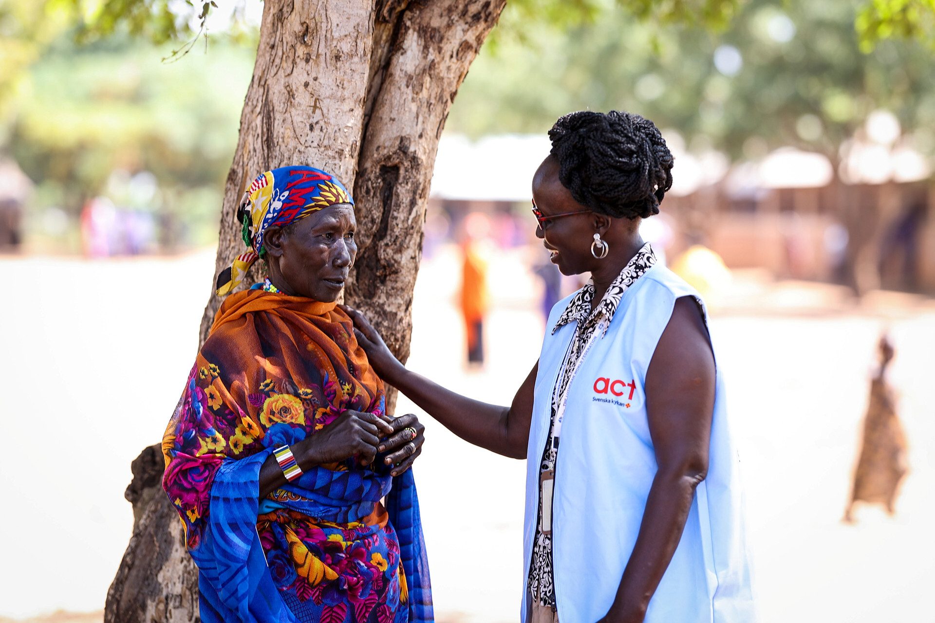 An aid worker from ACT Svenska kyrkan speaking with an older woman in traditional attire outdoors.