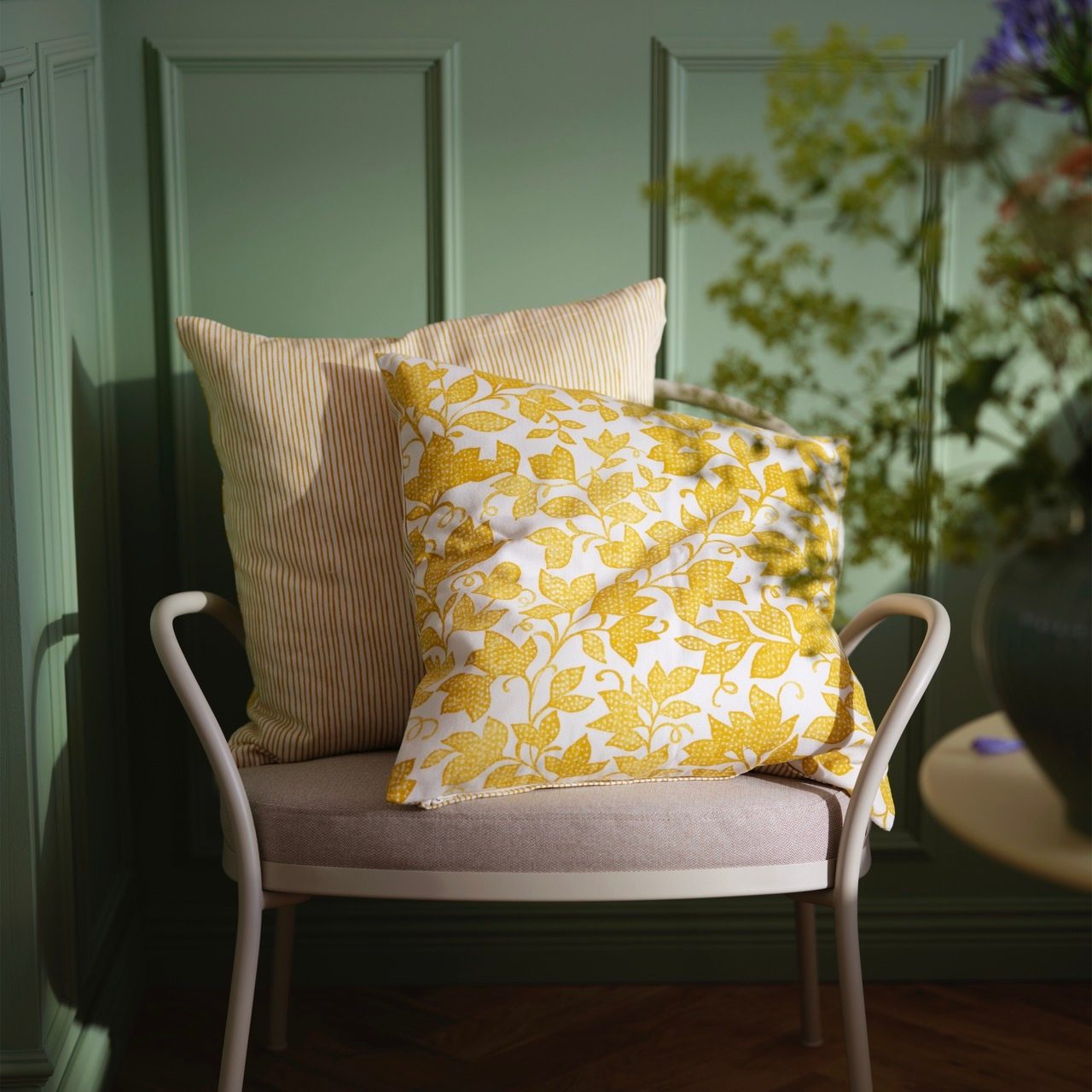 Two yellow patterned cushions on a beige chair, against a green paneled wall with sunlight.