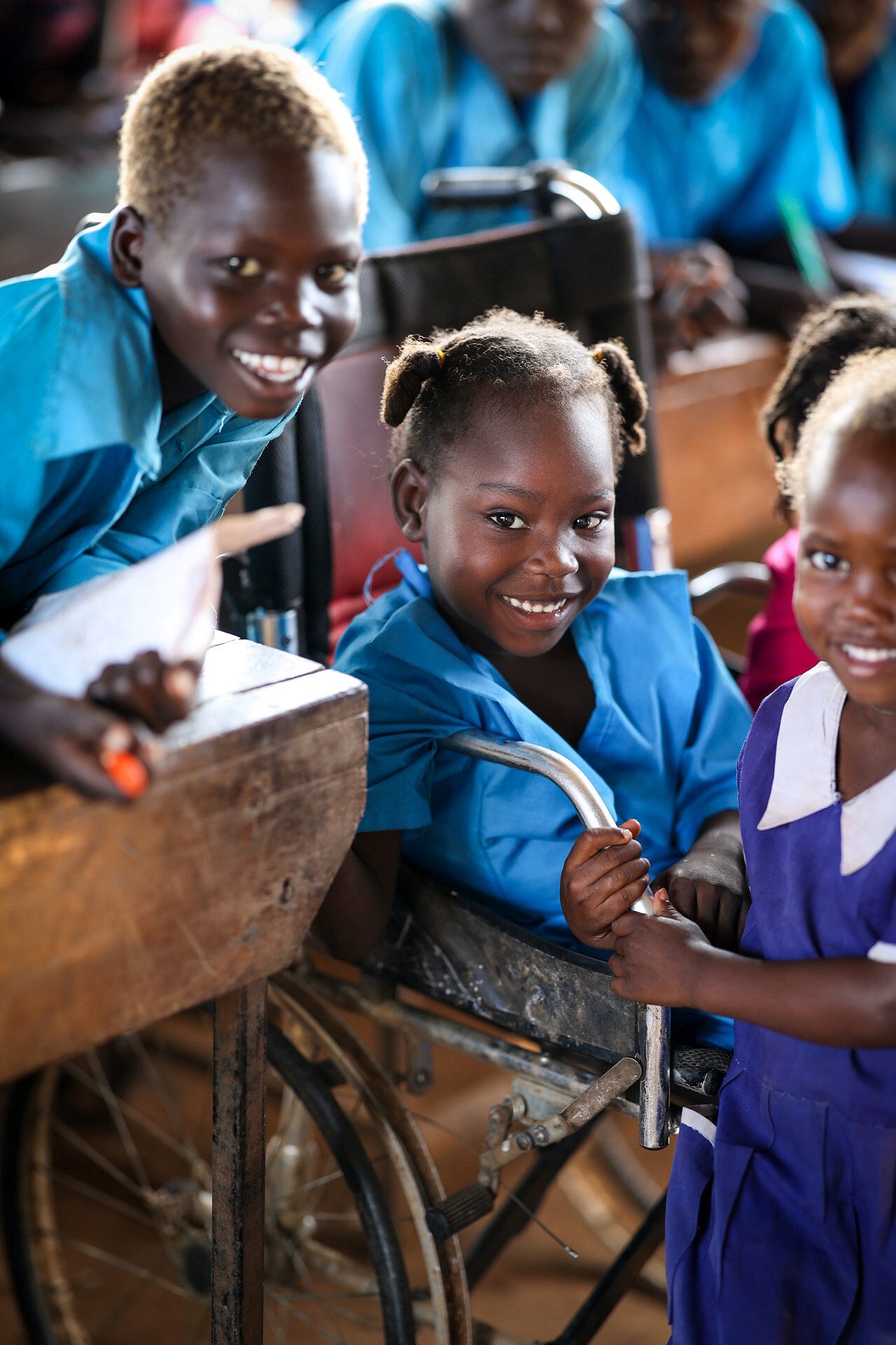 Three smiling children in a classroom, one girl in a wheelchair.