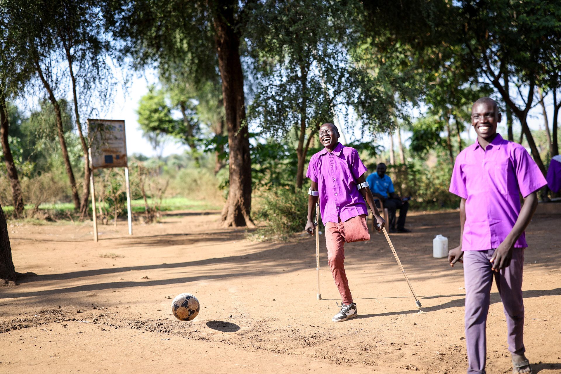 Joyful young amputee on crutches kicks soccer ball, smiling friend nearby on dirt field.