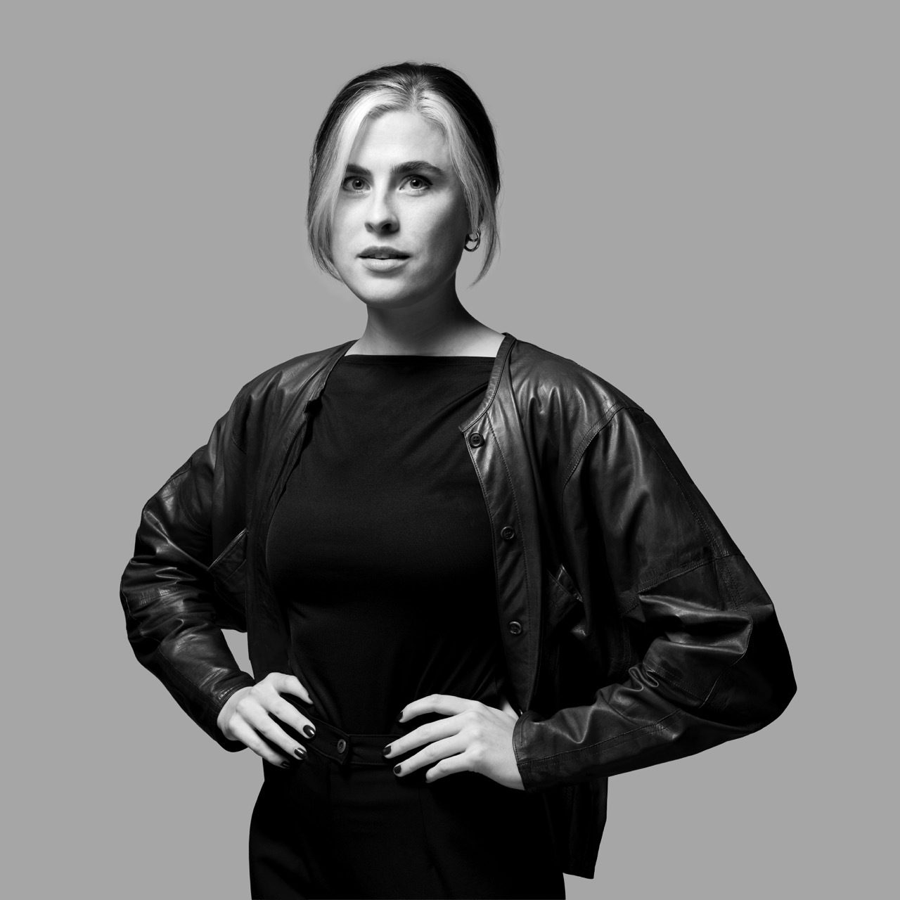 Black and white portrait of a woman with two-toned hair, black leather jacket, and hands on hips.