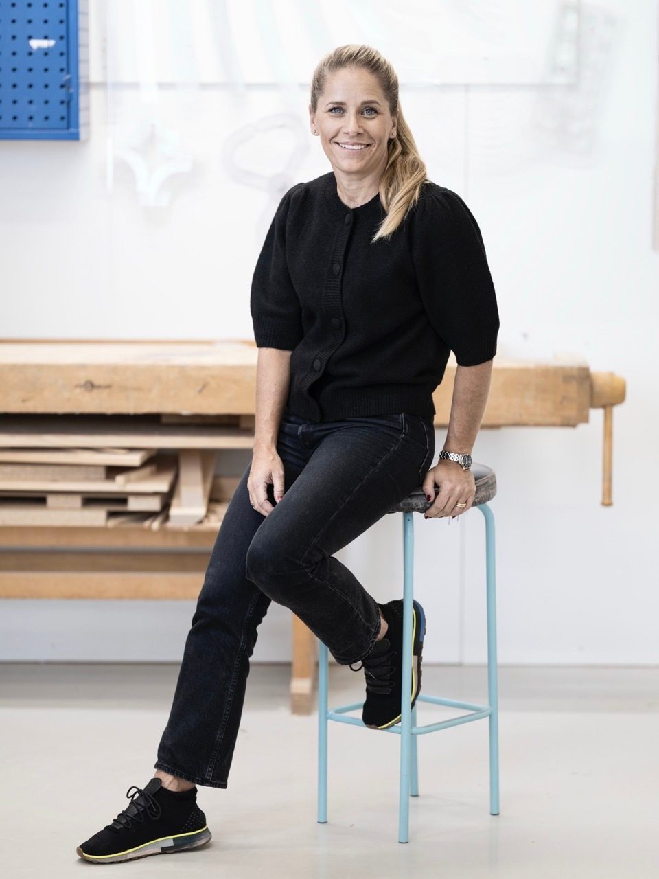A smiling blonde woman in a black cardigan and jeans sits on a blue stool in a workshop.