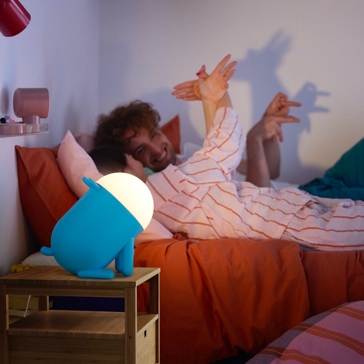 Smiling person in bed making shadow puppets, with a glowing blue lamp nearby.