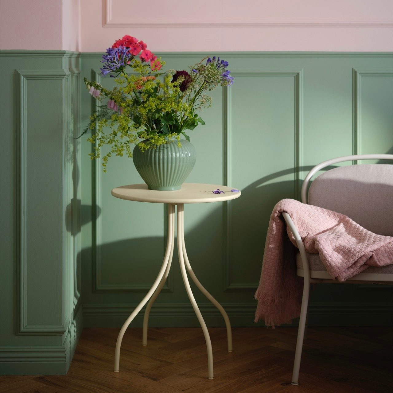 Vase of colorful flowers on a small table next to a chair with a pink blanket, green and pink room.