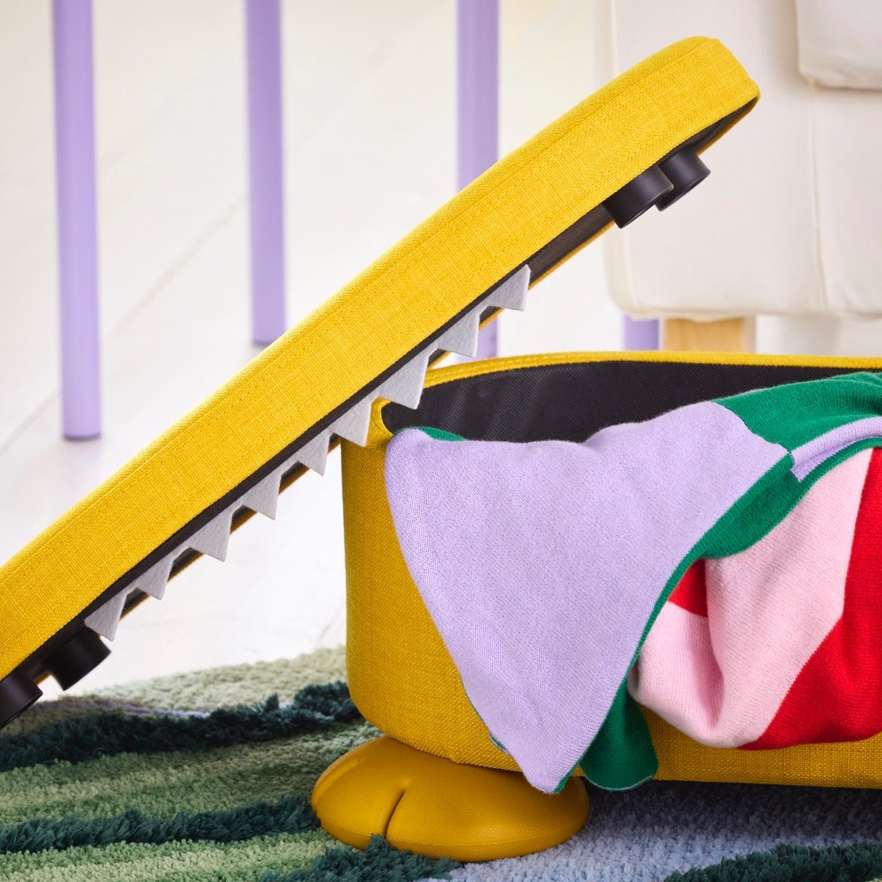 Yellow crocodile storage box, mouth open with teeth, colorful clothes inside, on a green rug.