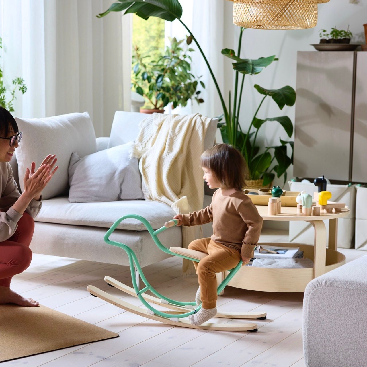 Mother claps while child rides a green rocking horse in a cozy living room.