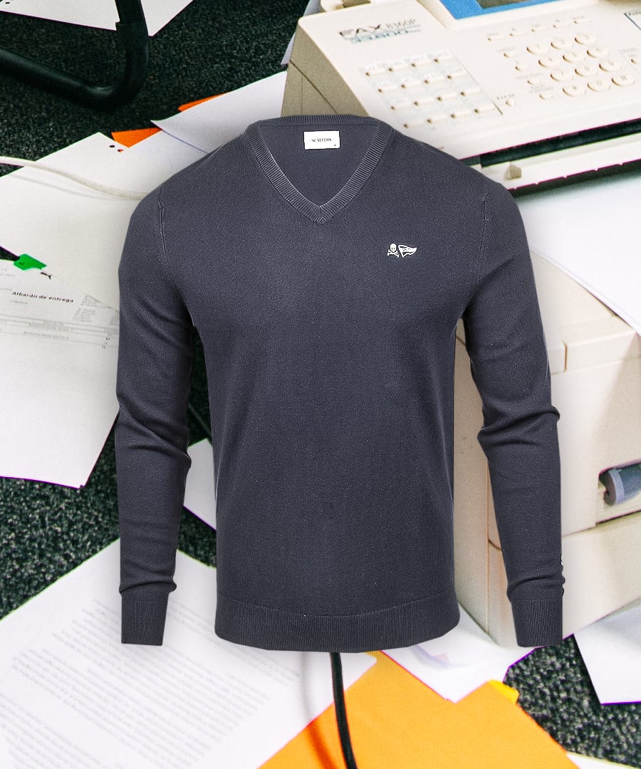 A dark gray V-neck sweater with a white embroidered logo, displayed against an office background.