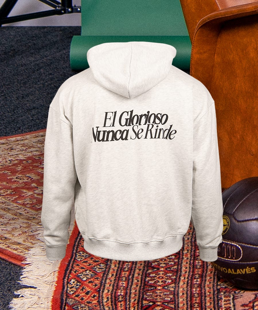 Back view of a light grey hoodie with "El Glorioso Nunca Se Rinde" text, on a patterned rug.
