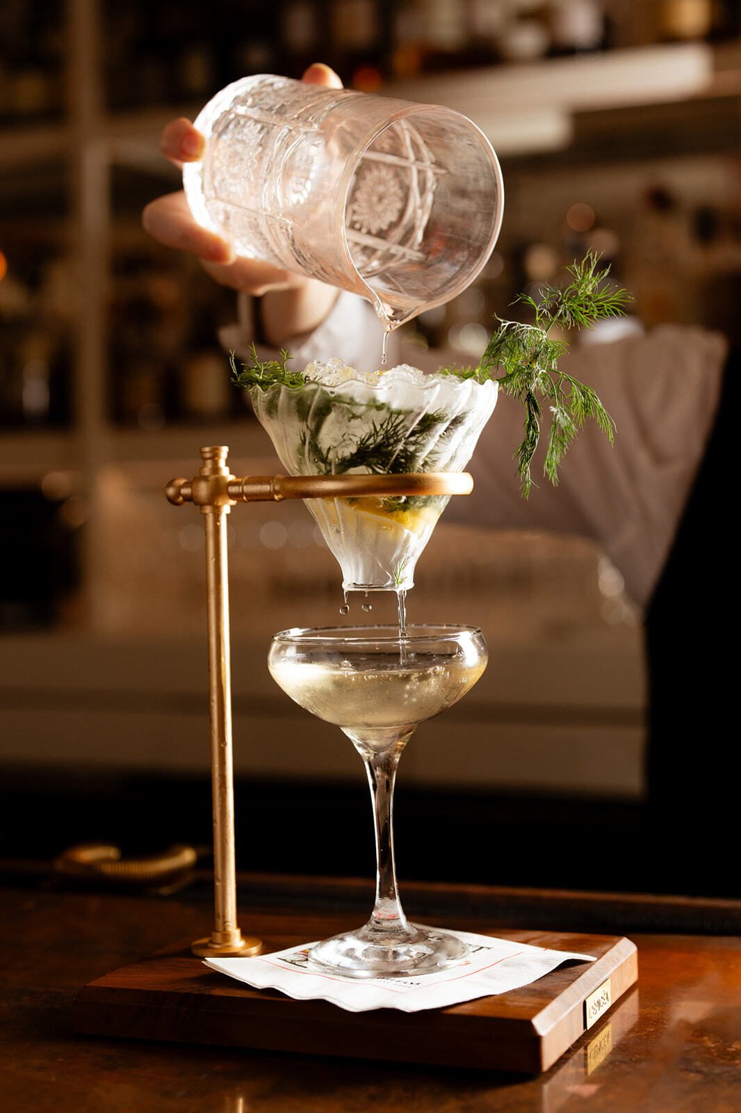 A bartender pours liquid from a patterned glass through a filter with ice and herbs into a cocktail coupe.