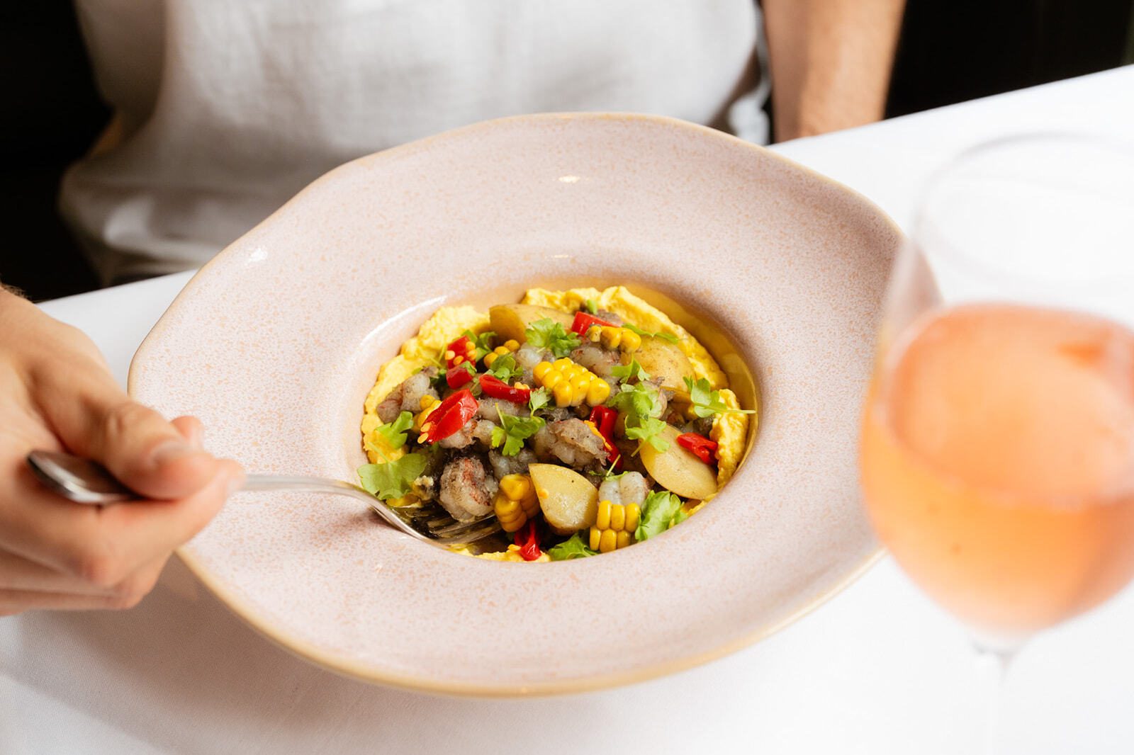 A person eating a colorful seafood and vegetable dish with a fork, served alongside a glass of rosé wine.