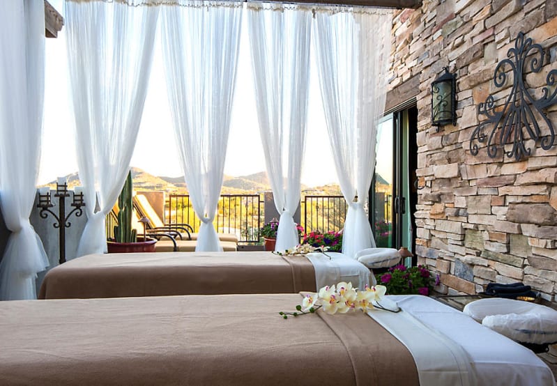 Outdoor spa with two massage beds, sheer white curtains, mountains, and a stone wall.