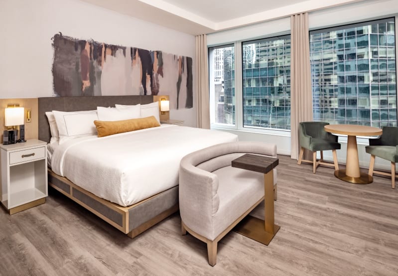 Bright modern hotel room with a bed, city view windows, and a small sitting area.