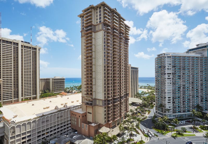 Tall brown hotel building with many balconies in a coastal city, other buildings, and ocean view.