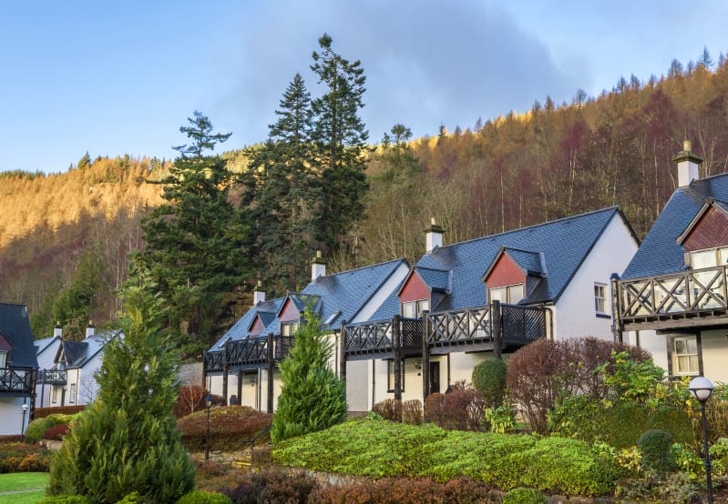 White houses with dark roofs and balconies, surrounded by green trees and a sunlit wooded hill.