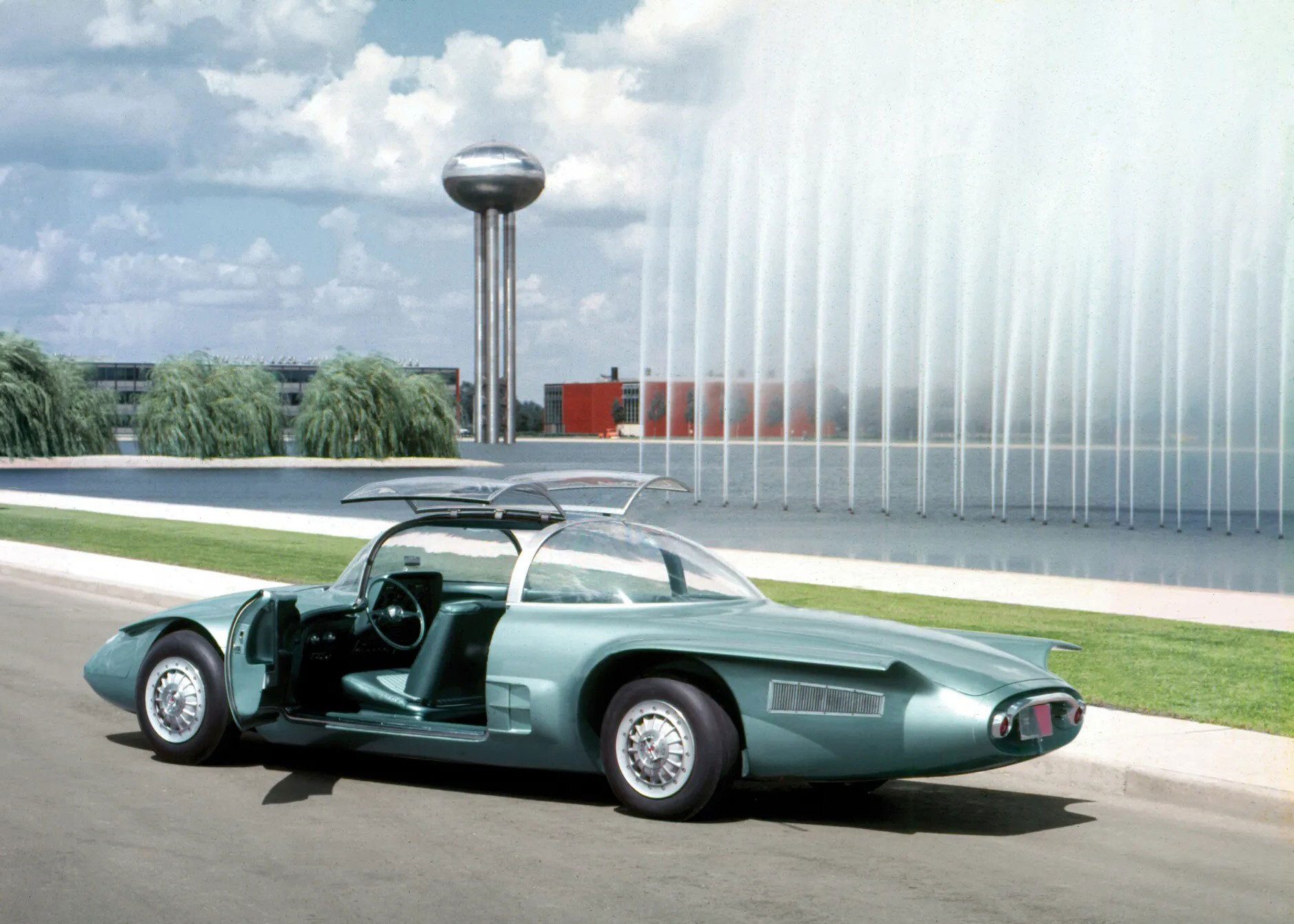 Futuristic teal concept car with gullwing doors open, parked next to a large fountain and a modern water tower.