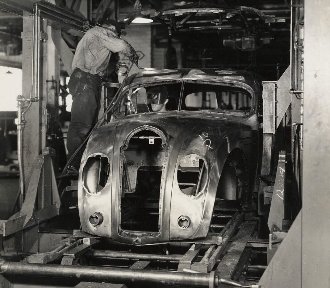 Two workers welding an unfinished vintage car body on an assembly line.