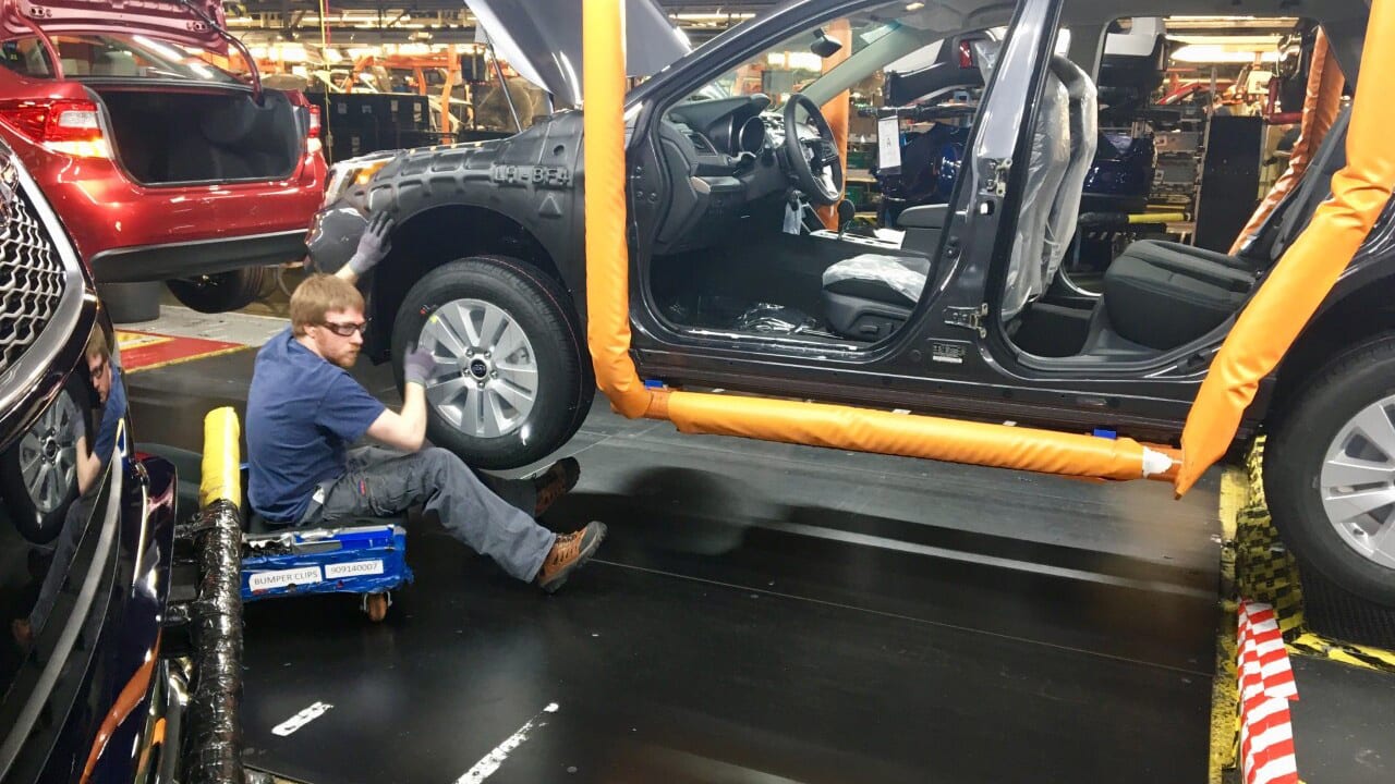 Worker on a creeper assembling a wheel on a car on an assembly line with other vehicles.