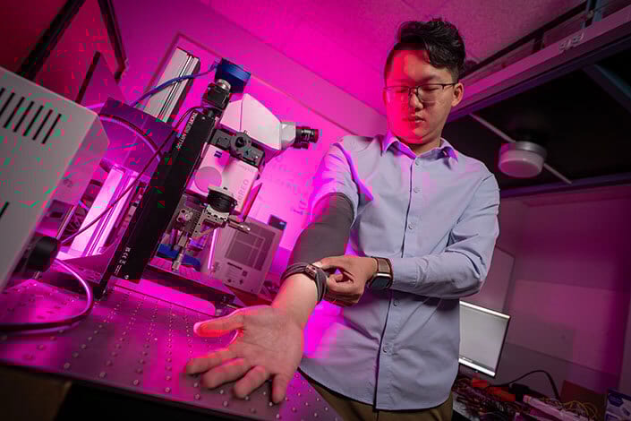 Man in a pink-lit lab adjusts a device on his forearm, near scientific equipment.