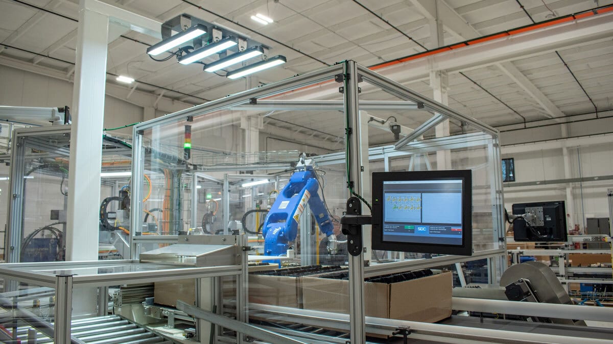 Blue industrial robot in a guarded factory cell with conveyors and a control screen.