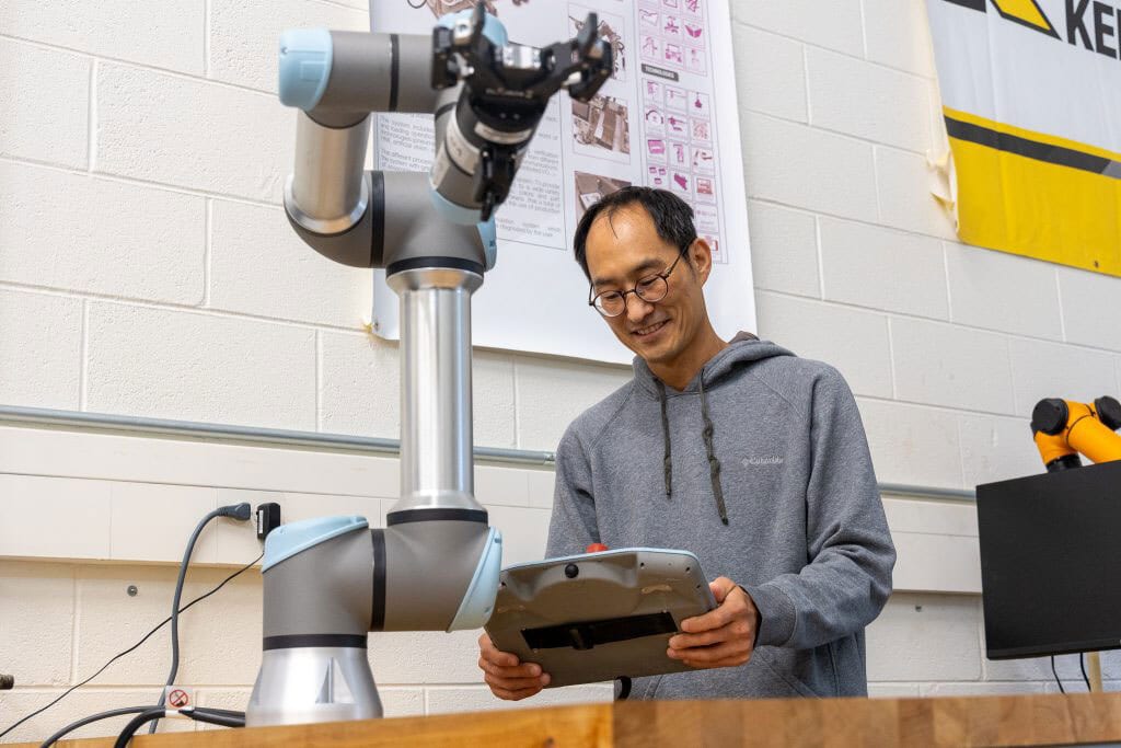 Smiling man in glasses operating a robotic arm with a tablet in a lab.