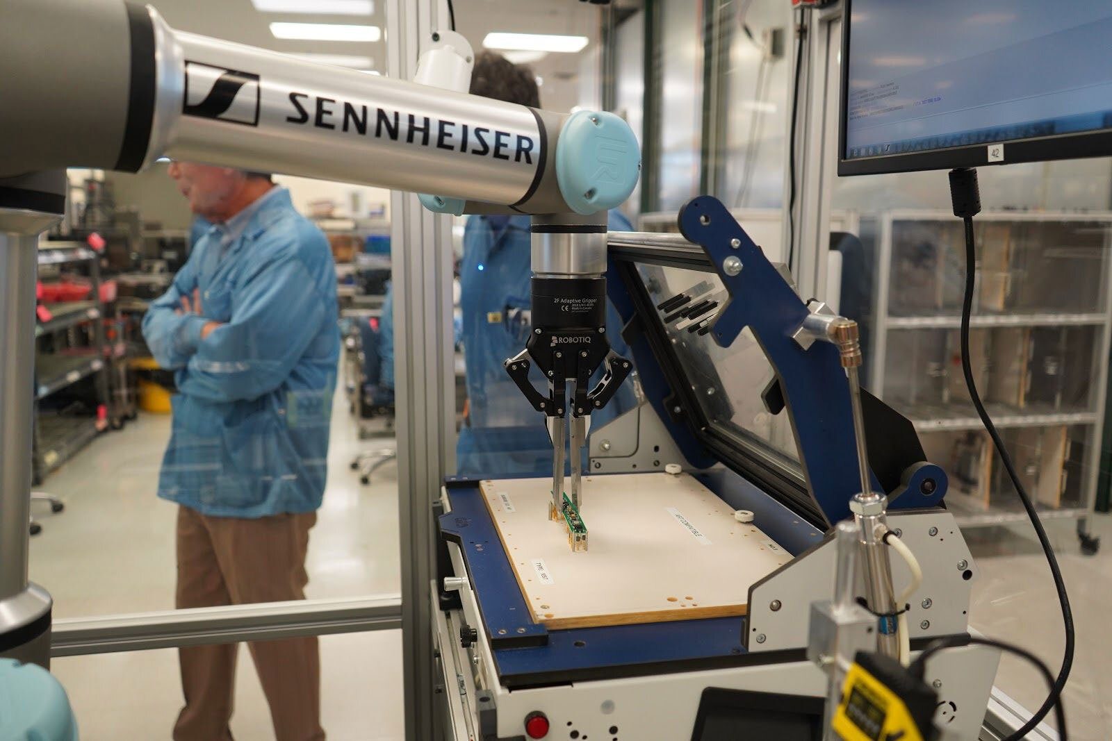 A Sennheiser robot arm grips a circuit board in a factory setting, with workers in background.