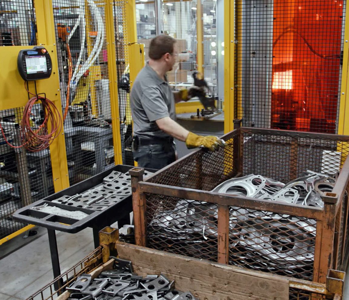 Factory worker loads metal parts into a machine with glowing orange light visible through mesh.
