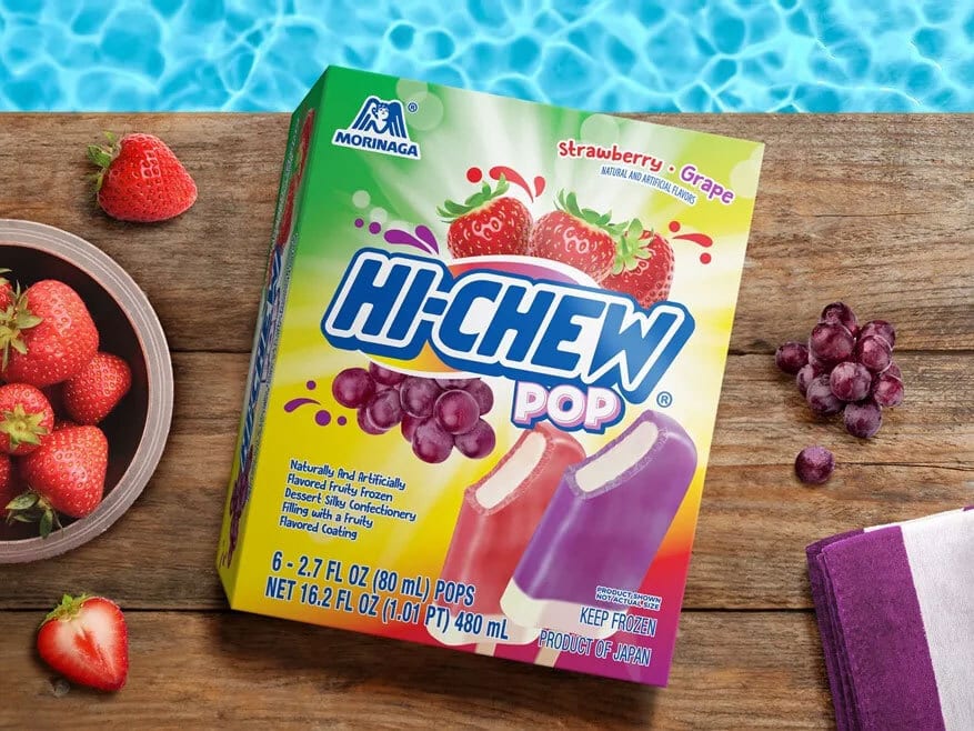 Hi-Chew Pop box (strawberry-grape flavor) next to a bowl of strawberries and scattered grapes on a wooden deck by a pool.