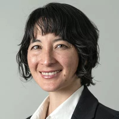 A smiling woman with short dark hair, bangs, and freckles, wearing a white shirt and dark jacket.