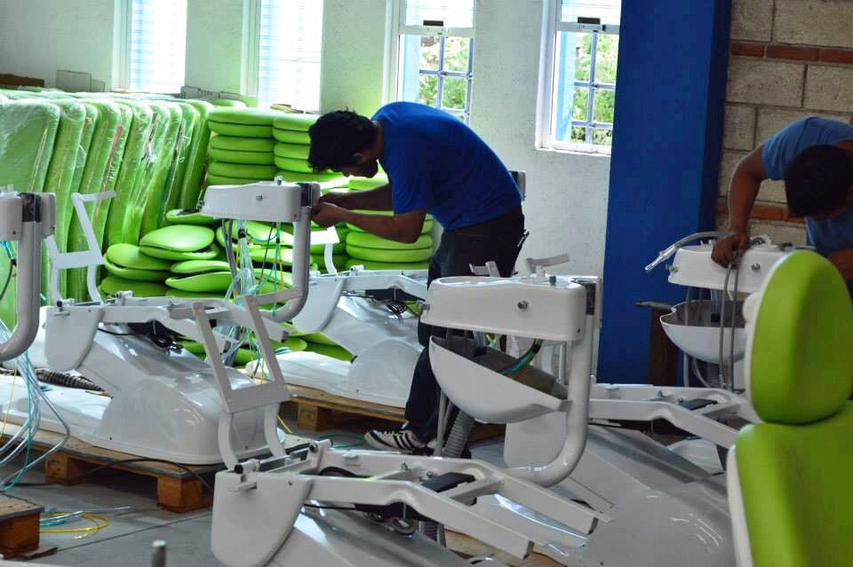Workers assemble white dental chairs with bright green cushions in a busy factory workshop.