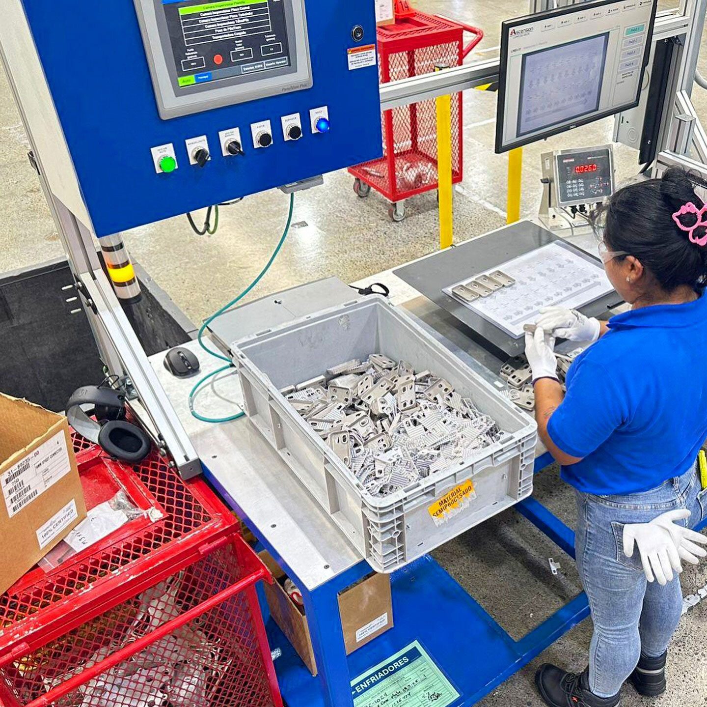A factory worker inspecting small metal parts at a workstation with machinery and screens.