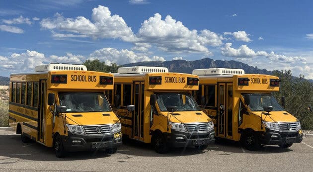 Three yellow school buses parked outdoors with mountains and clouds.