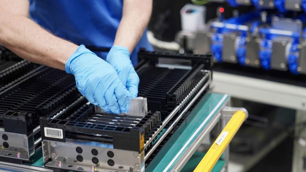 Hands in blue gloves place components into black racks on an assembly line.
