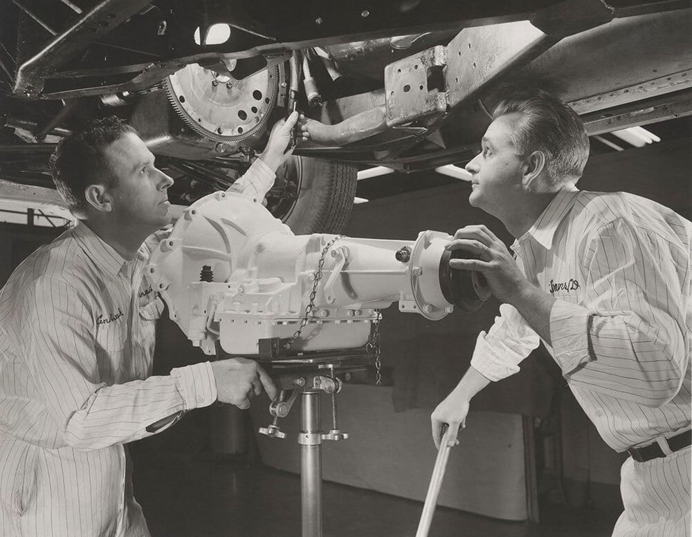 Two mechanics in striped uniforms work together to install a large car transmission or engine assembly, looking up at their work.