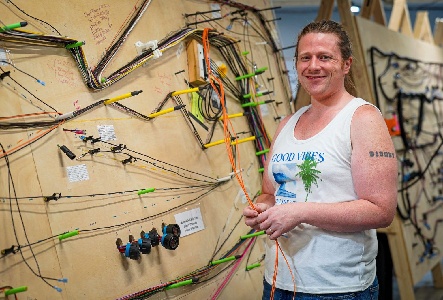 A smiling person in a tank top stands beside a complex electrical wiring harness board, holding orange wires.