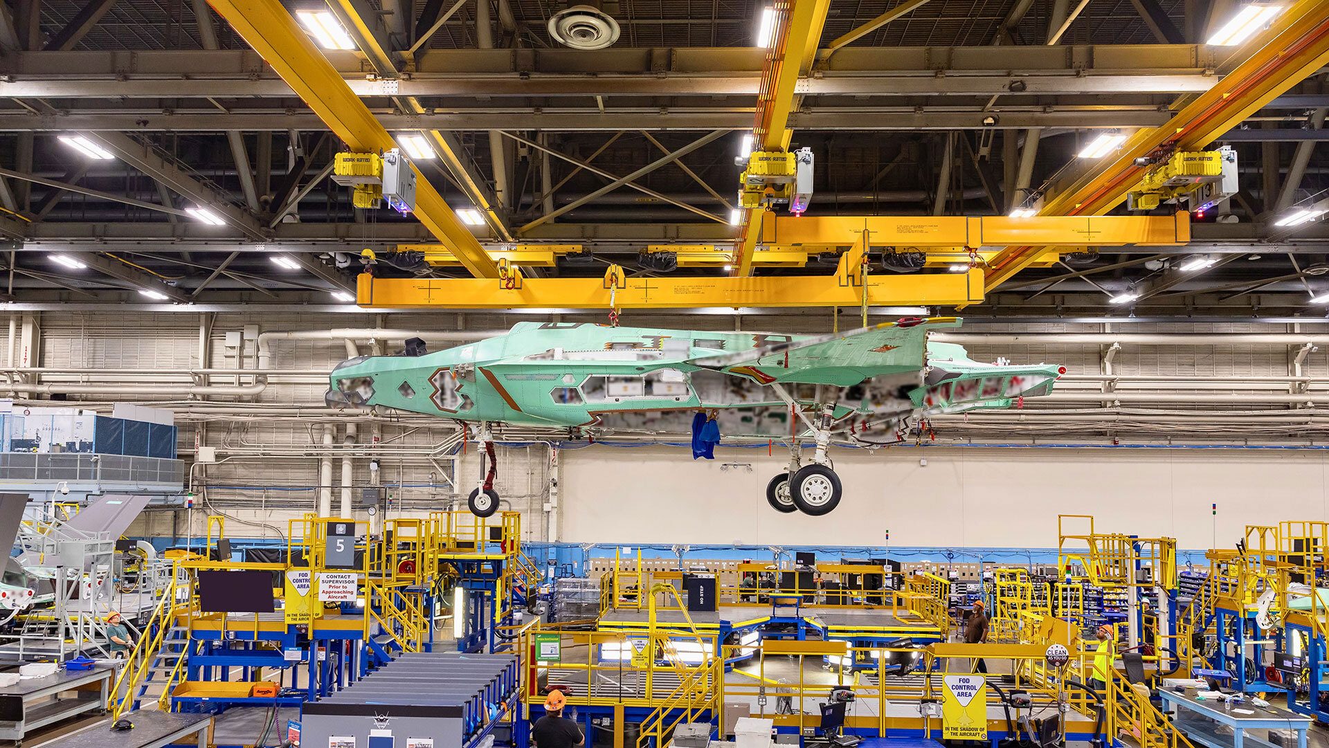 Light green fighter jet fuselage under assembly, suspended by cranes in a factory.