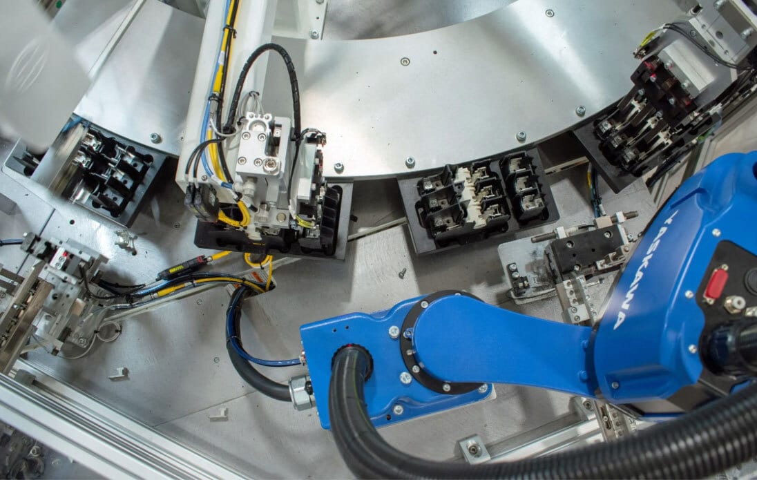 A blue Yaskawa robot arm within an industrial assembly system, surrounded by components, trays, and wiring.